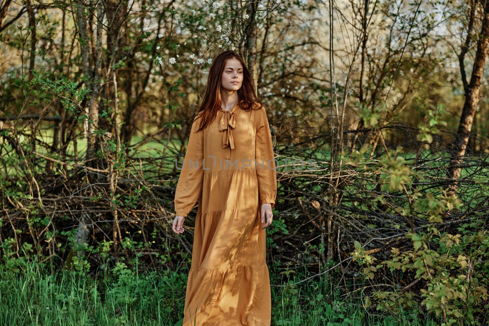 a beautiful woman walks in the shade near the trees, dressed in a long orange dress, enjoying the weather and the weekend. The theme of privacy with nature, horizontal photography on the street. High quality photo