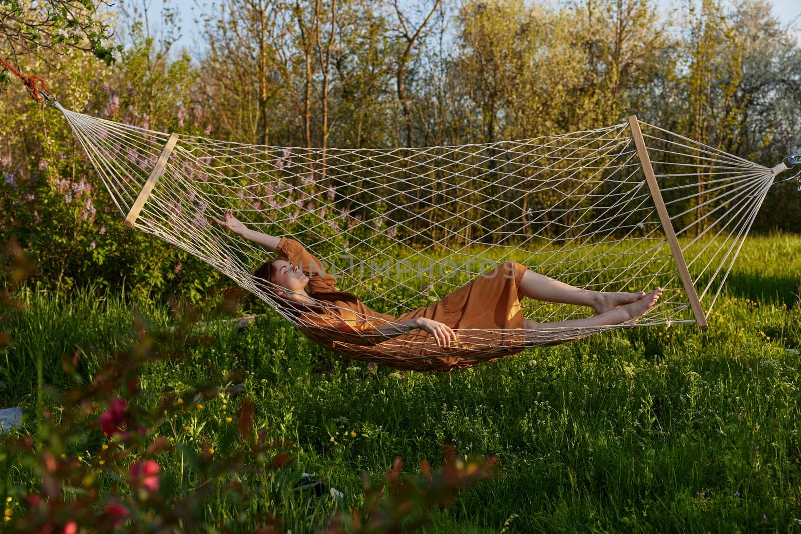 a happy woman in a long orange dress is relaxing in nature lying in a mesh hammock enjoying summer and vacation by Vichizh