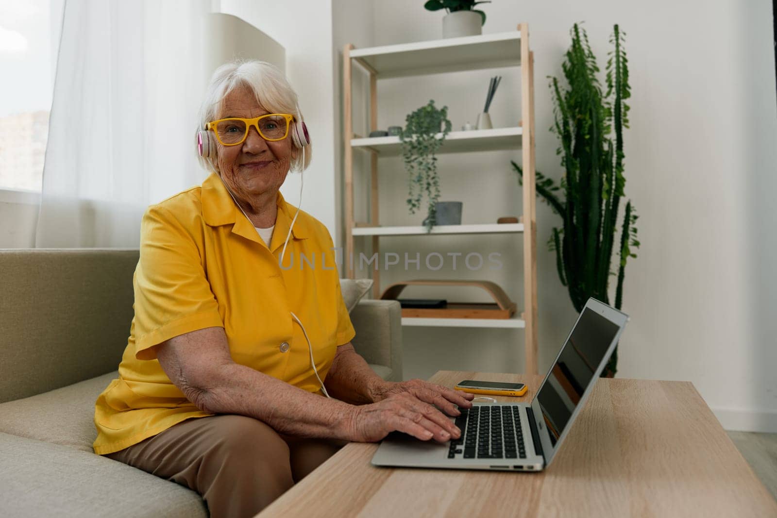 Happy elderly woman with a laptop typing with headphones sitting at home on the couch in a yellow shirt, bright modern interior, lifestyle online communication. High quality photo