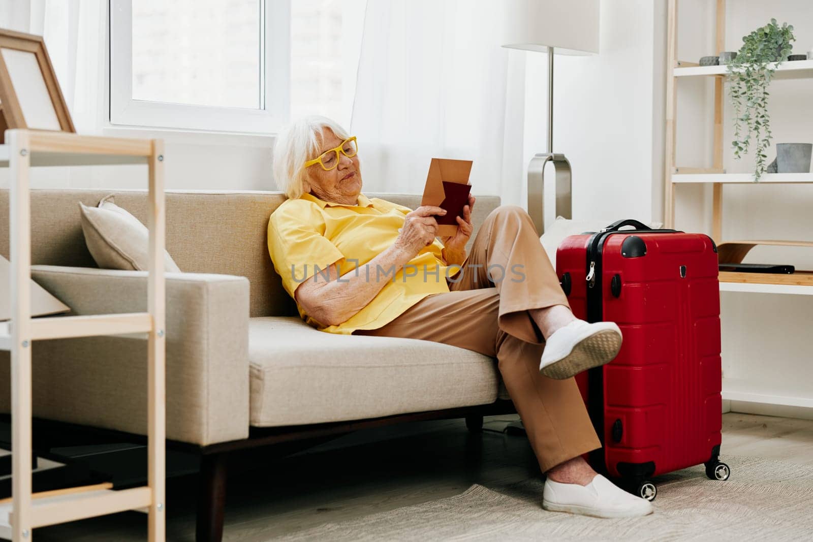 Happy senior woman with passport and travel ticket packed a red suitcase, vacation and health care. Smiling old woman joyfully sitting on the sofa before the trip raised her hands up in joy. High quality photo