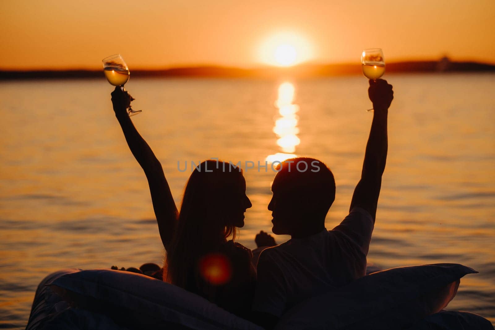 Silhouettes of a happy couple raising glasses on a summer evening near the sea at sunset.