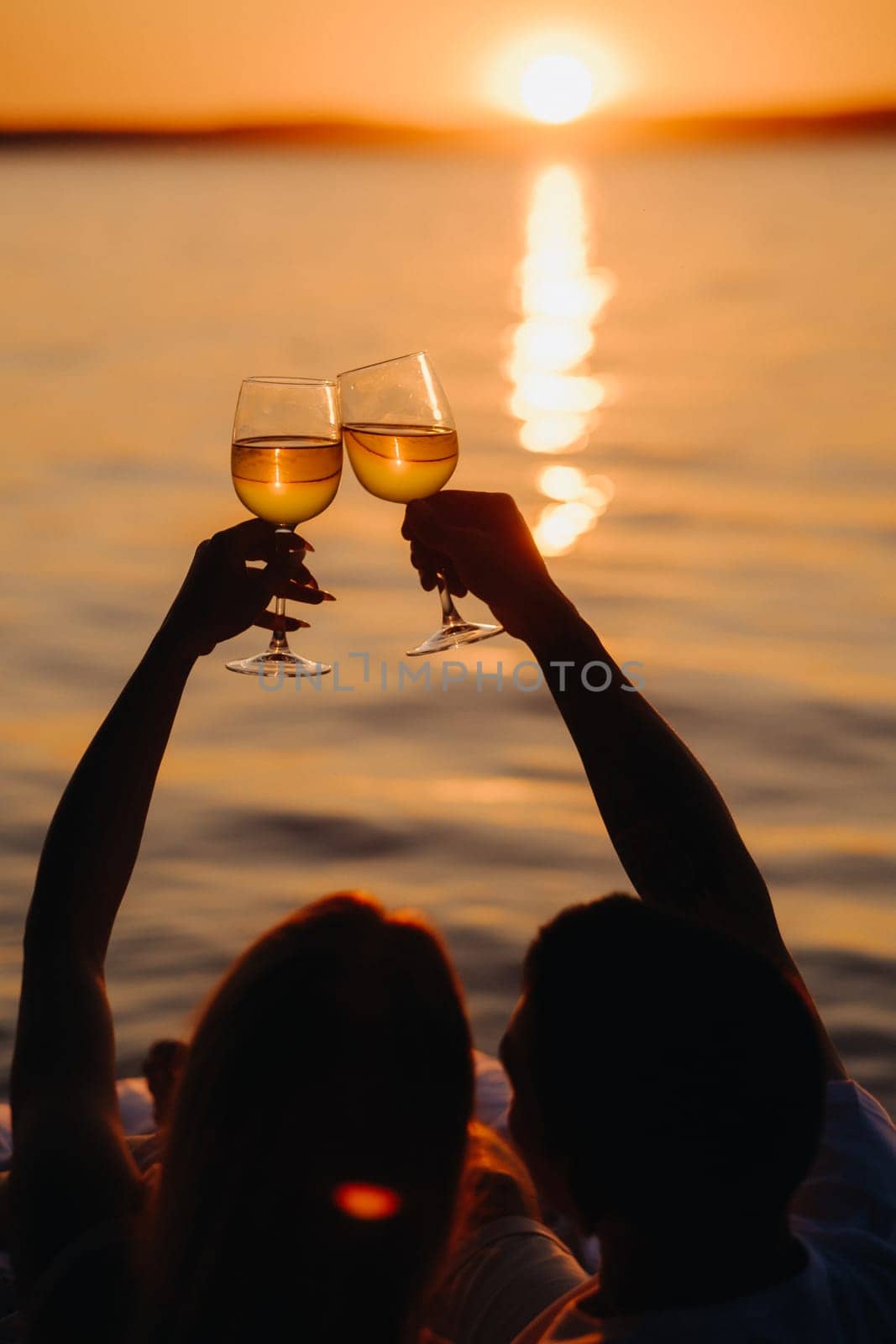 Silhouettes of a happy couple raising glasses on a summer evening near the sea at sunset.