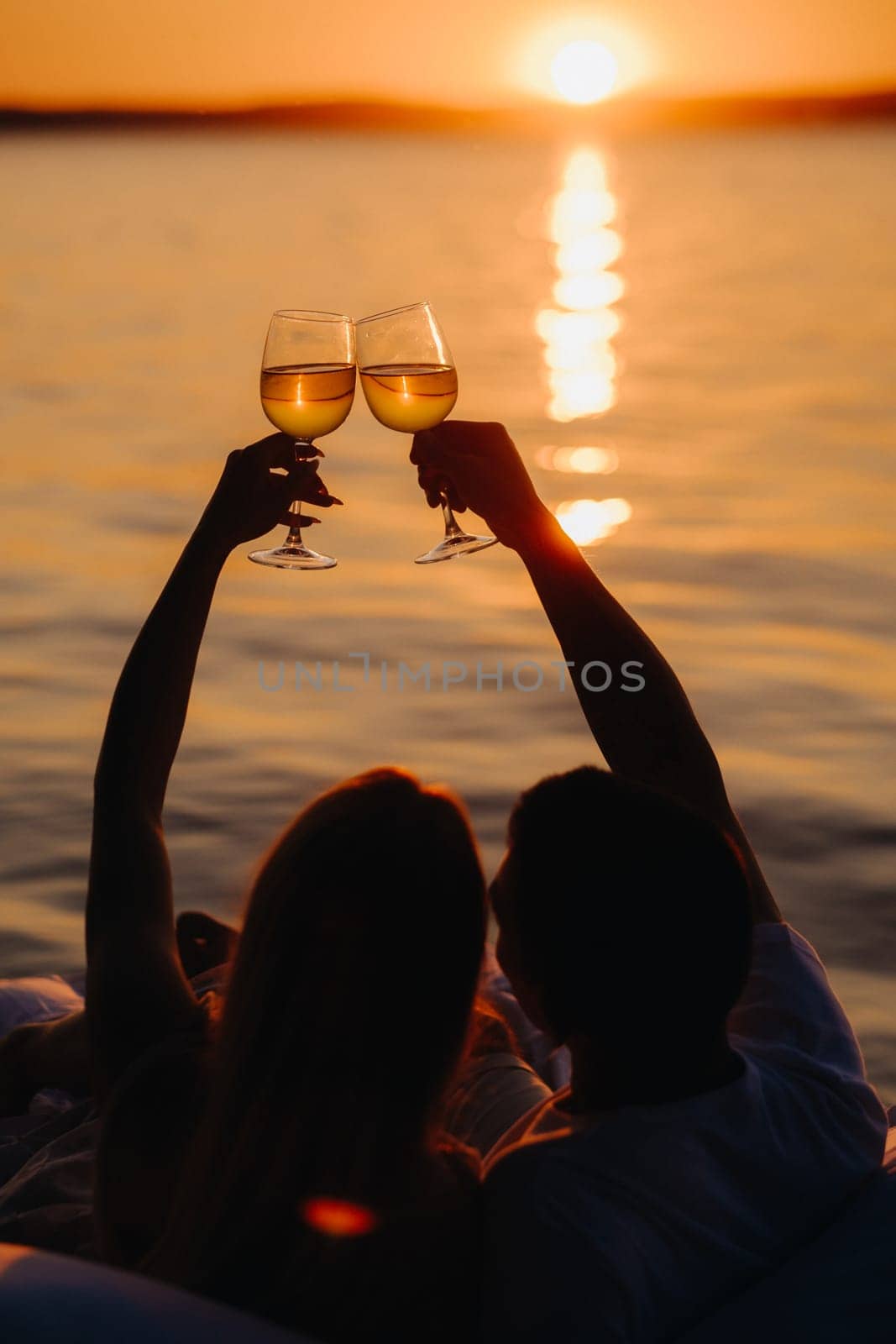 Silhouettes of a happy couple raising glasses on a summer evening near the sea at sunset.