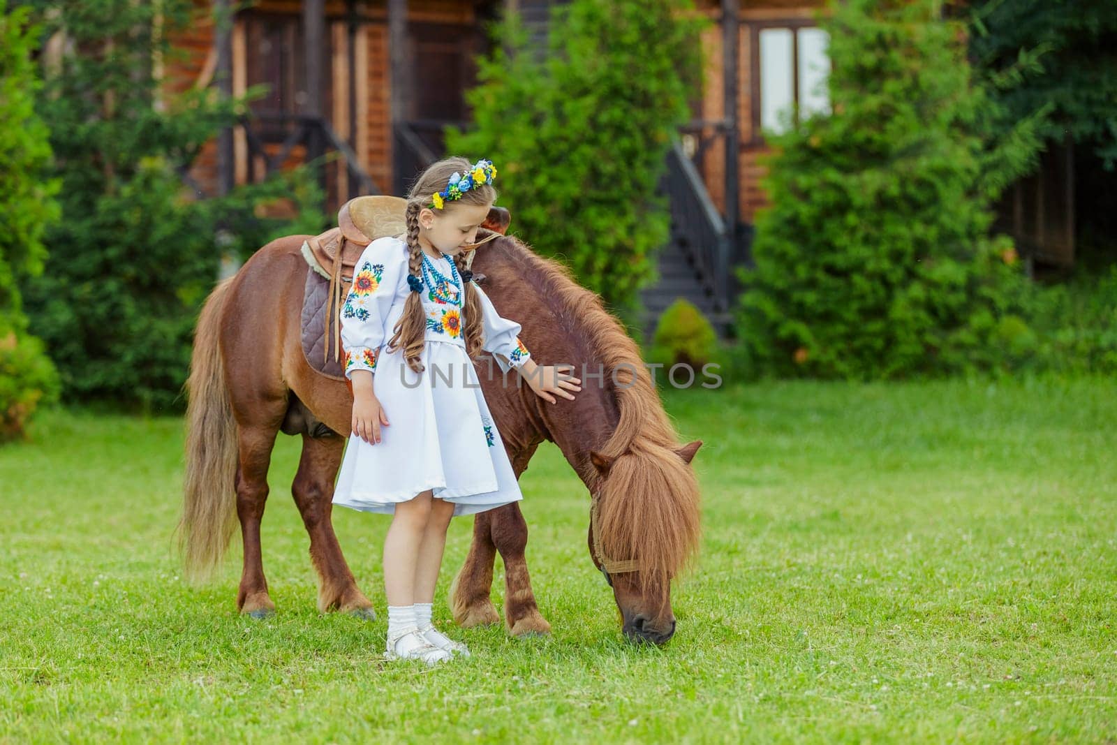 young girl in Ukrainian national dress strokes a pony that grazes on the lawn