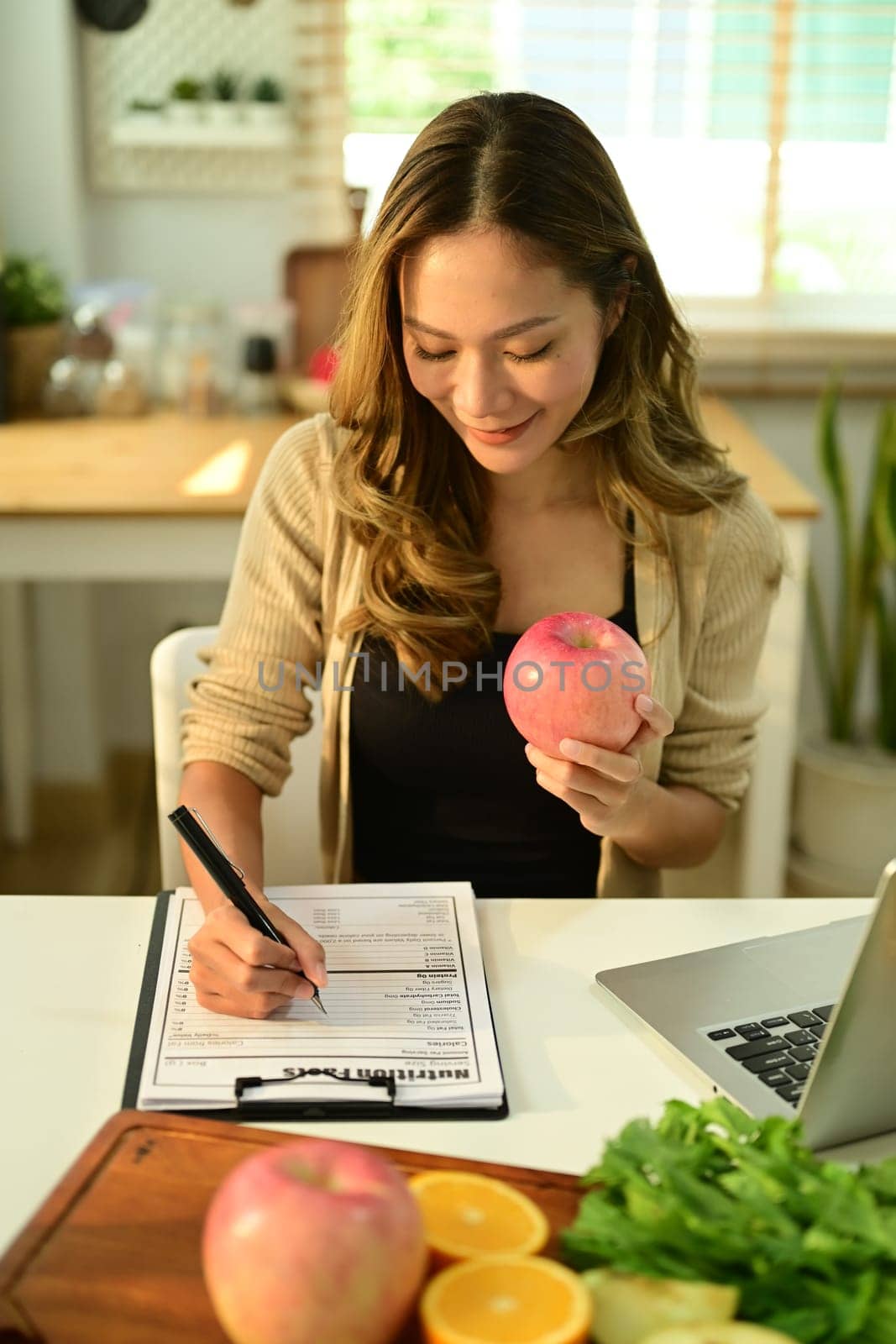 Charming young woman holding an apple and writing recipe at desk with fresh fruit. Dieting, right nutrition, healthy eating by prathanchorruangsak