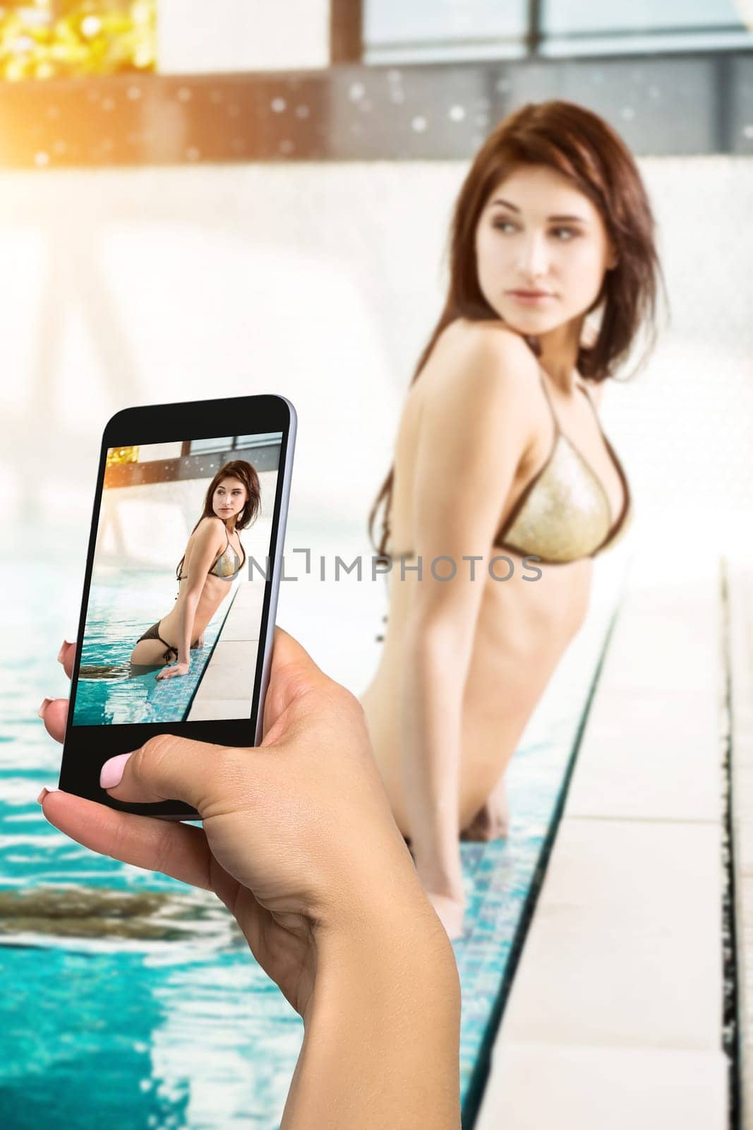 Closely image of female hands holding mobile phone with photo camera mode on the screen. Cropped image of beautiful long hair female model posing by the pool in wellness spa hotel resort.