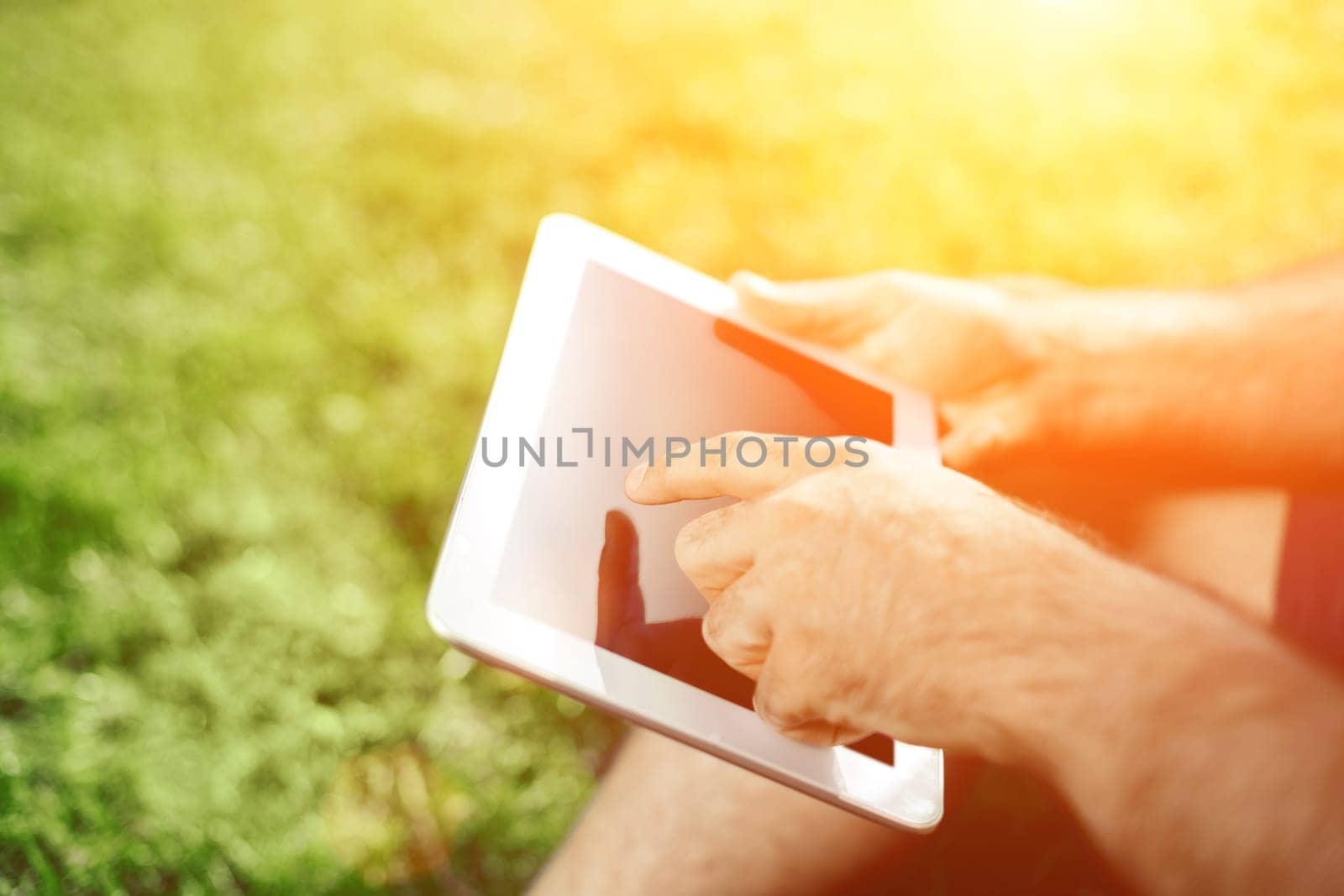 Close-up of casual dressed young man using modern digital tablet while sitting at the park, hipster man connecting social networking outside, sun light. Sun flare