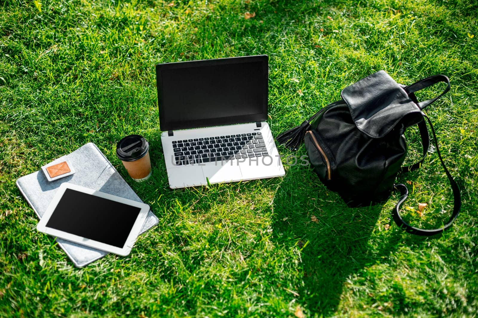 Laptop computer on green grass with coffee cup, bag and tablet in outdoor park. Copy space. Still life