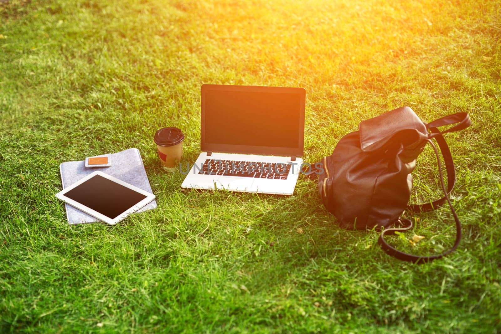 Laptop computer on green grass with coffee cup, bag and tablet in outdoor park. Copy space. Still life. Sun flare