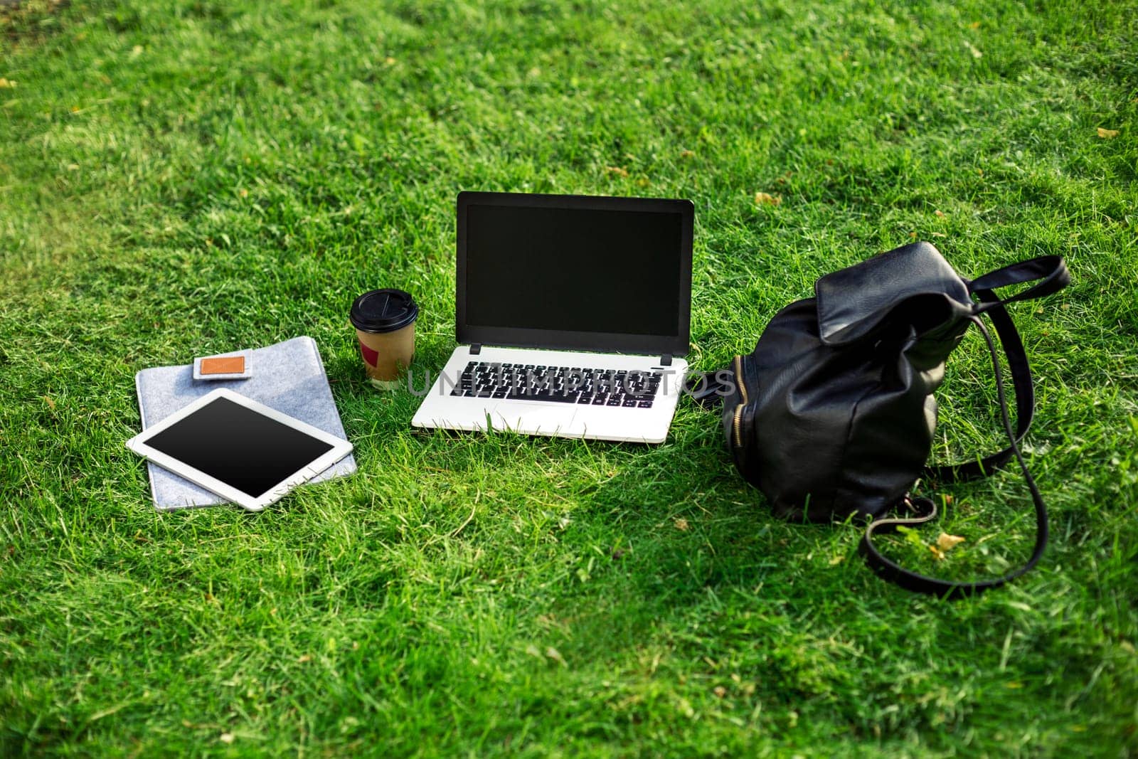 Laptop computer on green grass with coffee cup, bag and tablet in outdoor park. Copy space. Still life