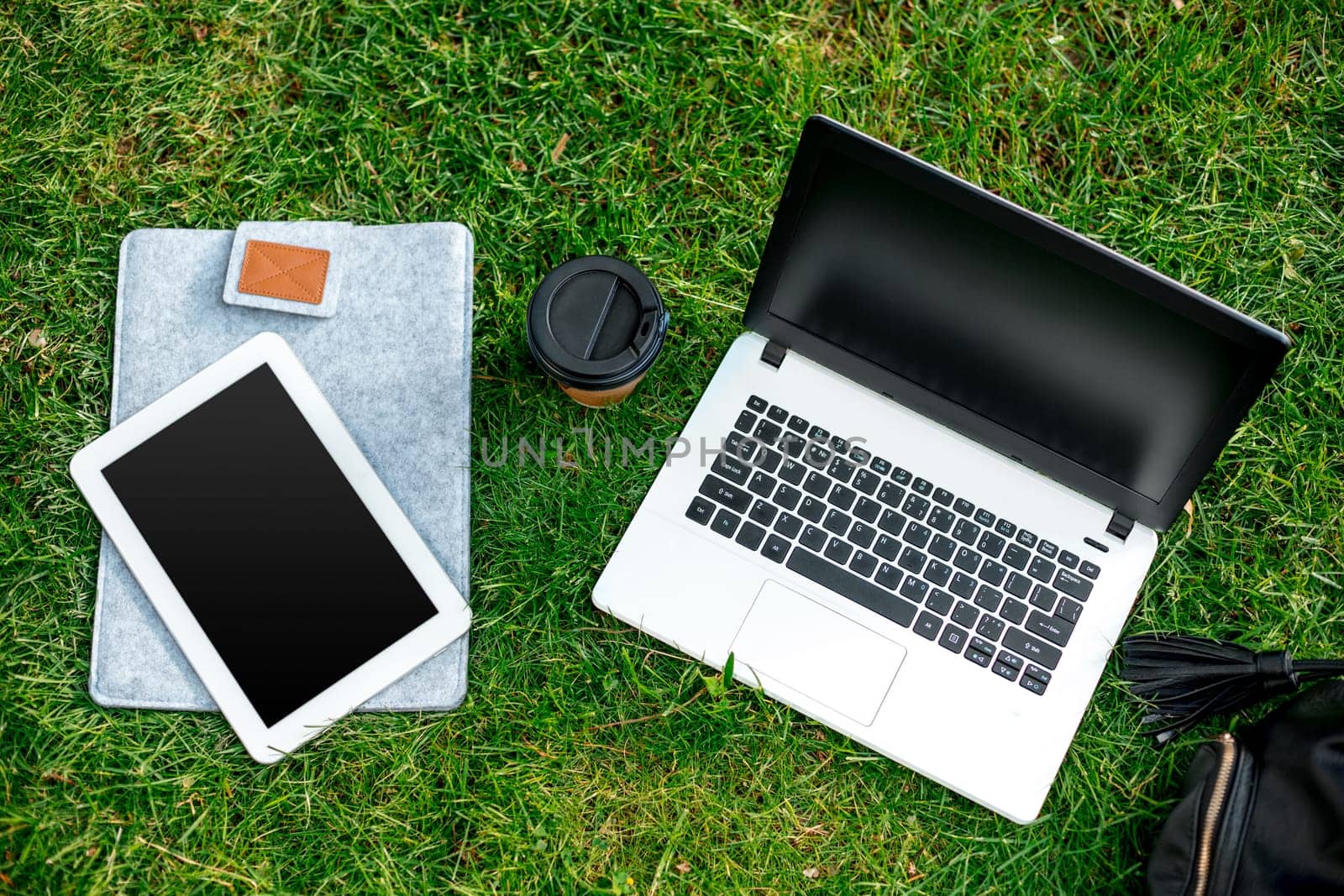 Laptop computer on green grass with coffee cup, bag and tablet in outdoor park. Copy space. Still life