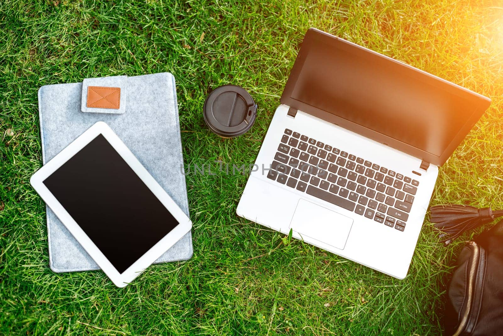 Laptop computer on green grass with coffee cup, bag and tablet in outdoor park. Copy space. Still life. Sun flare