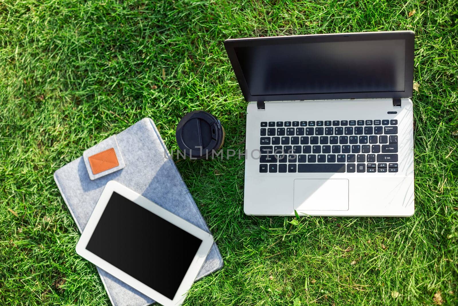 Laptop computer on green grass with coffee cup and tablet in outdoor park. Copy space. Still life