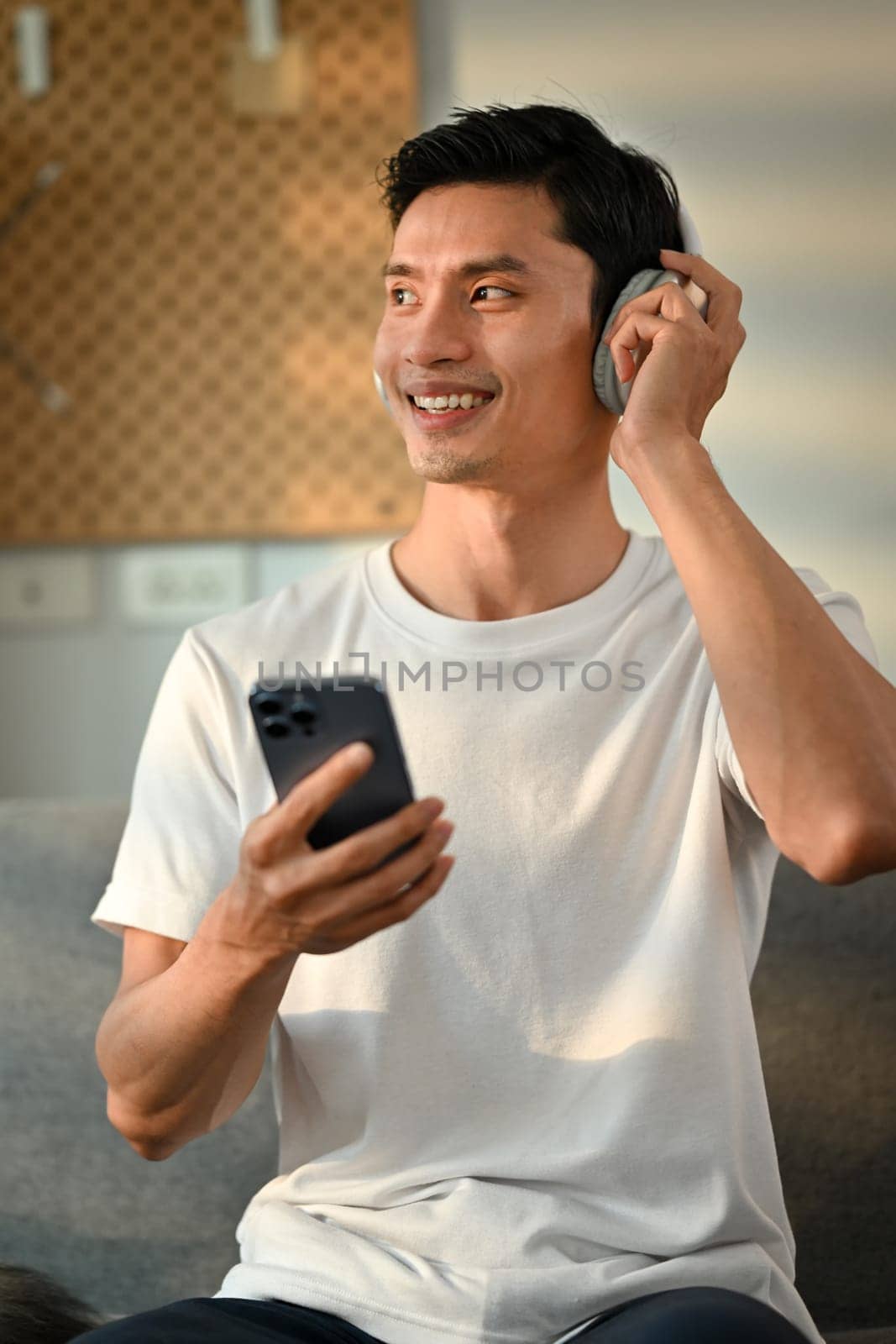 Happy millennial asian man enjoying listening to favorite music online on headphone and using smartphone in living room.