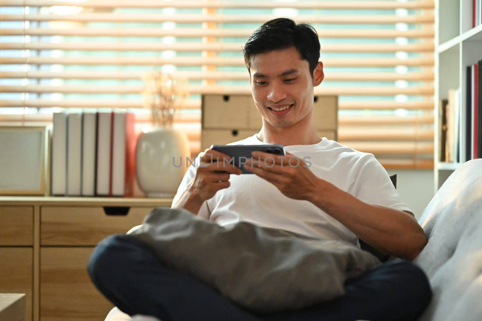 Relaxed asian man in casual wears watching video or playing game on smart phone, sitting on couch at home.
