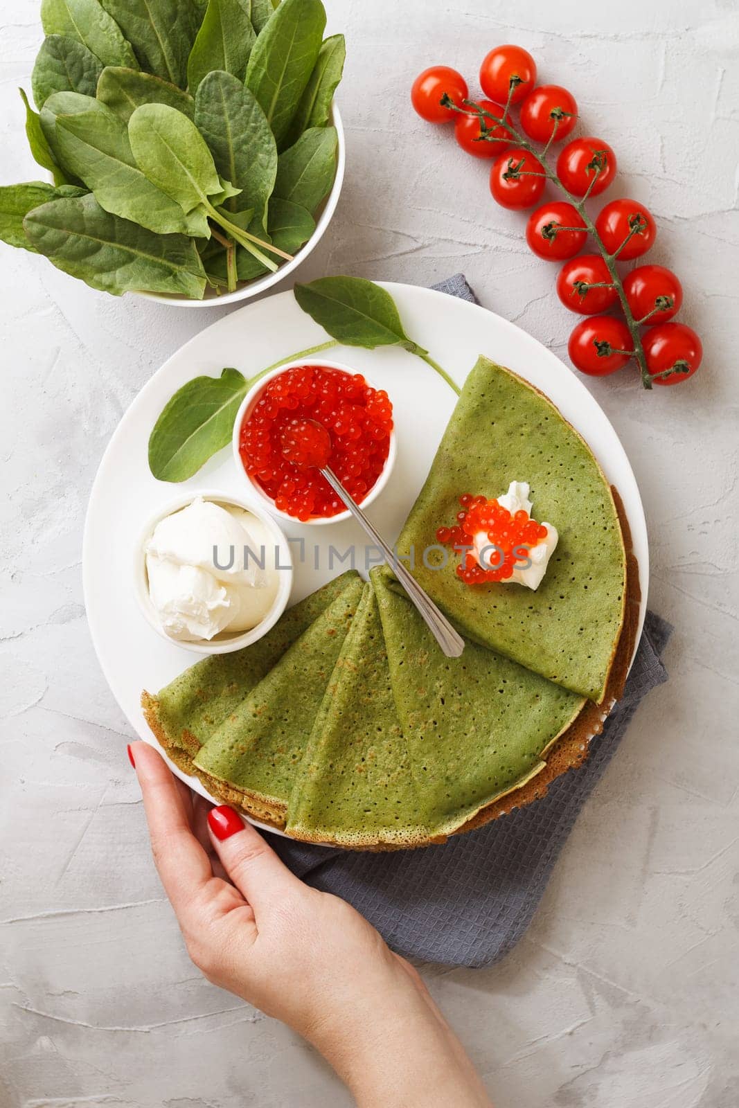 A woman's hand serves a plate of spinach pancakes with soft cheese, caviar and cherry tomatoes on a gray background with textiles. vertical photo by lara29