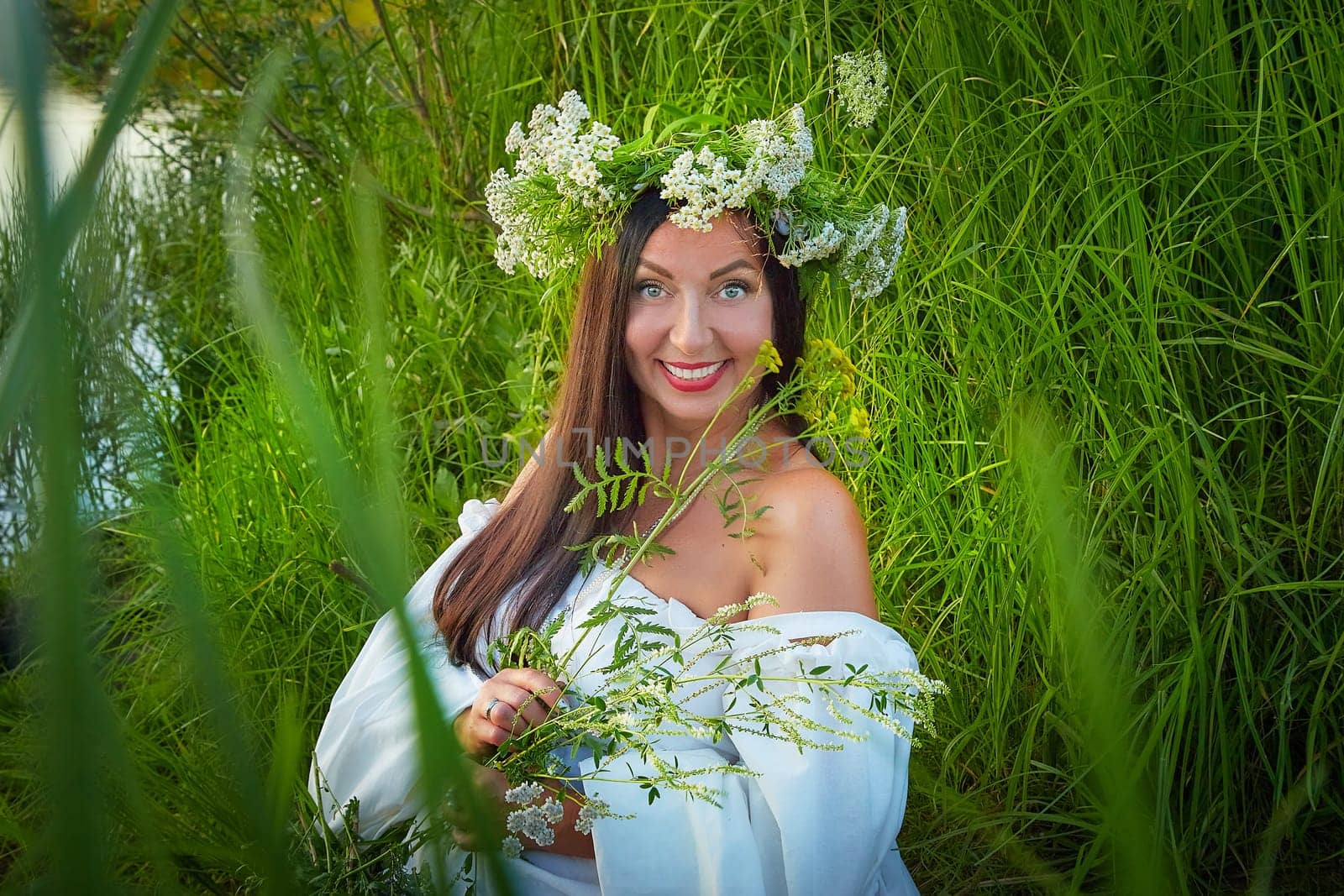 Adult mature brunette woman in a white dress, sundress and a wreath of flowers in summer by the water of river or lake in evening at sunset. Celebration of the Slavic pagan holiday of Ivan Kupala