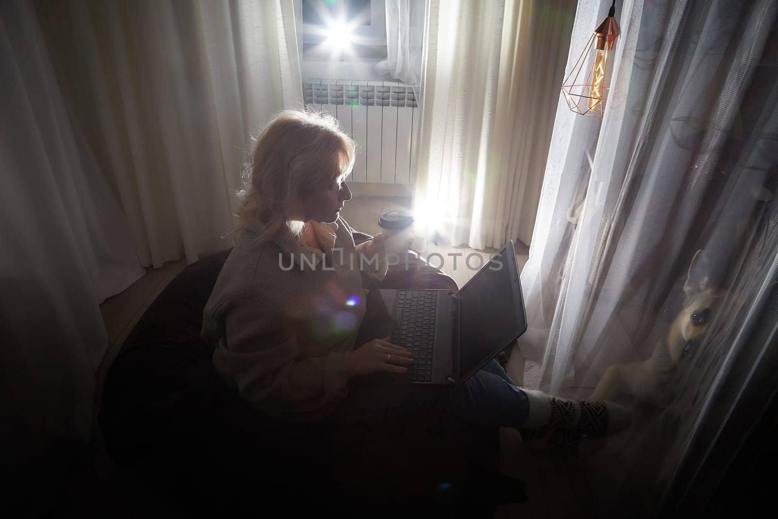 Silhouette and shadow of Adult mature woman of 40-60 years with laptop, notebook, computer in warm sweater in dark calm cozy evening atmosphere room. Interior with curtains and small linght