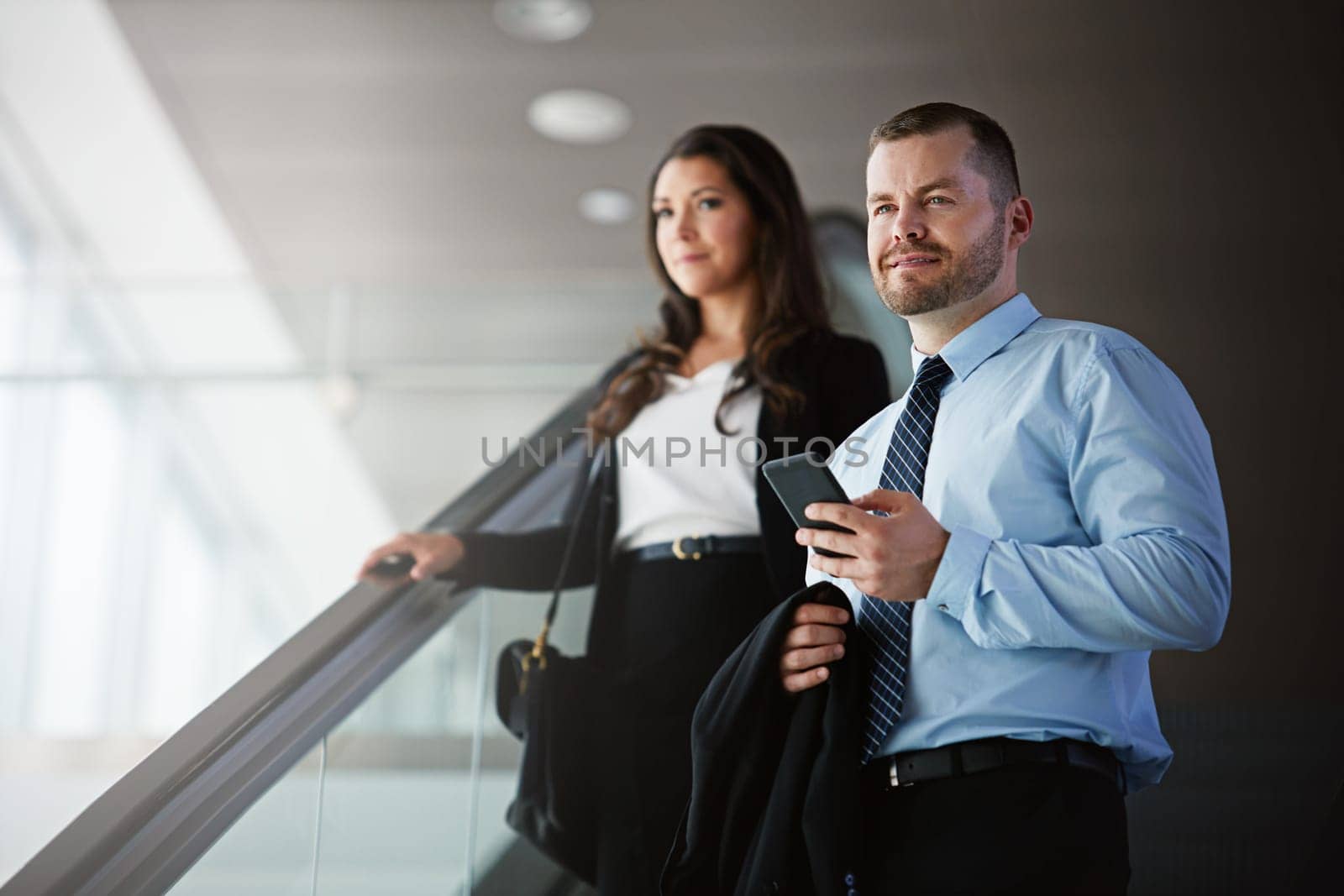 Business travel - moving to where the money is. a businessman and businesswoman traveling down an escalator in an airport
