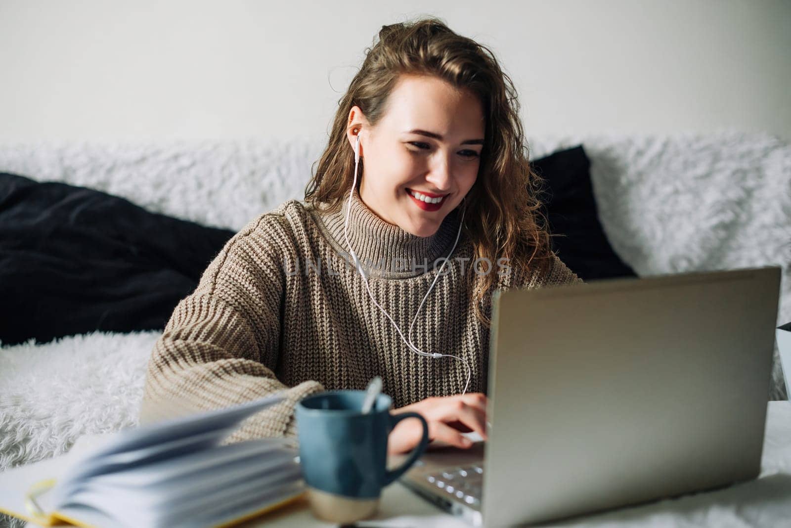 Student video chatting with friends, taking a break from studying at home. Young woman in headphones sitting at kitchen table with laptop and coffee, smiling while chatting with friends on video call.