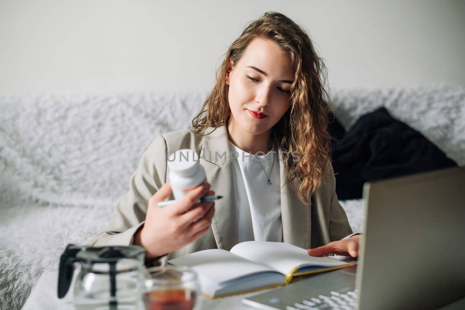Nutritionist writing article about effects of supplement deficiency on human health, working on laptop at kitchen table with bottle of vitamins nearby.