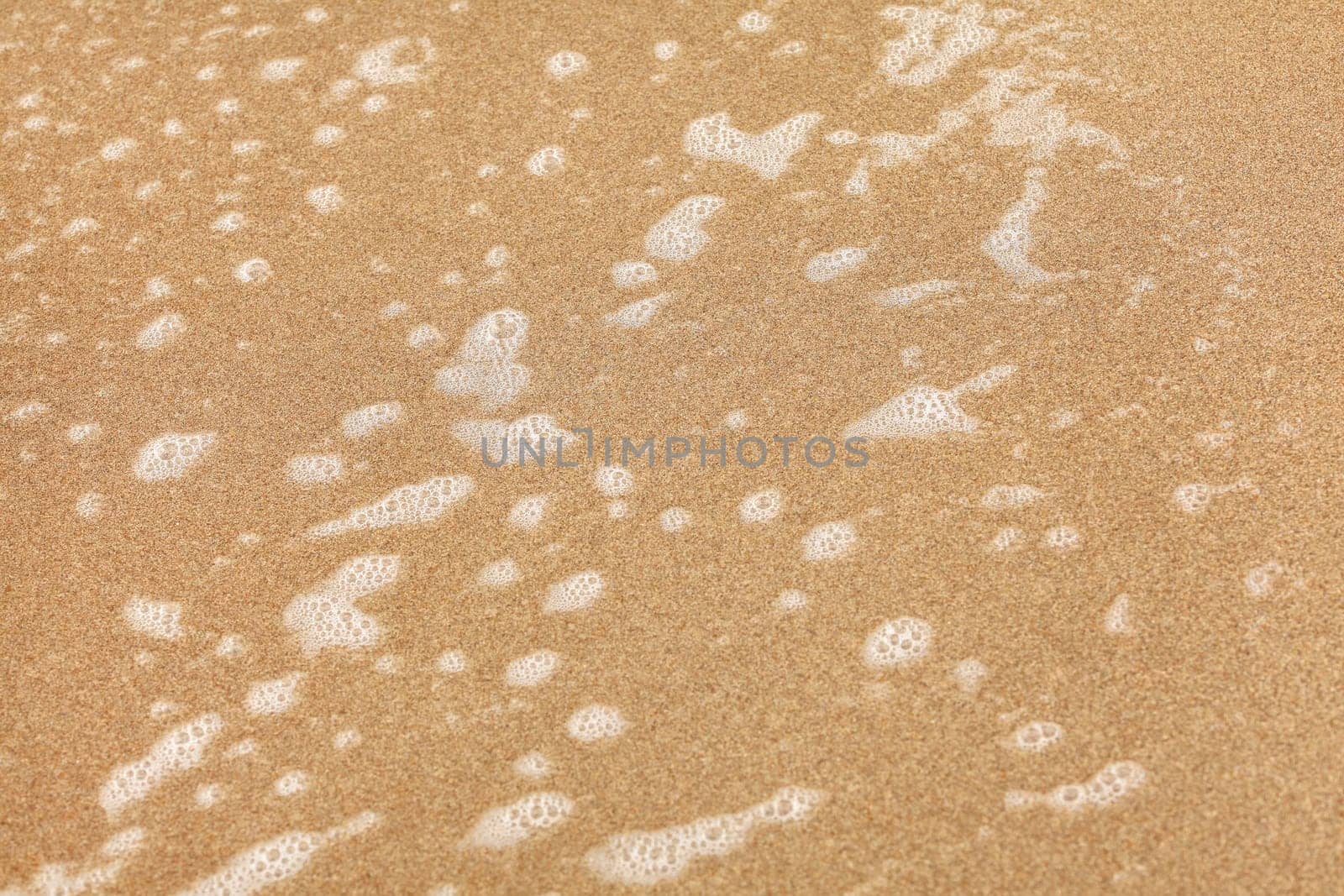 Spots of white foam on beach sand wet from ocean water. Abstract sea / marine background.