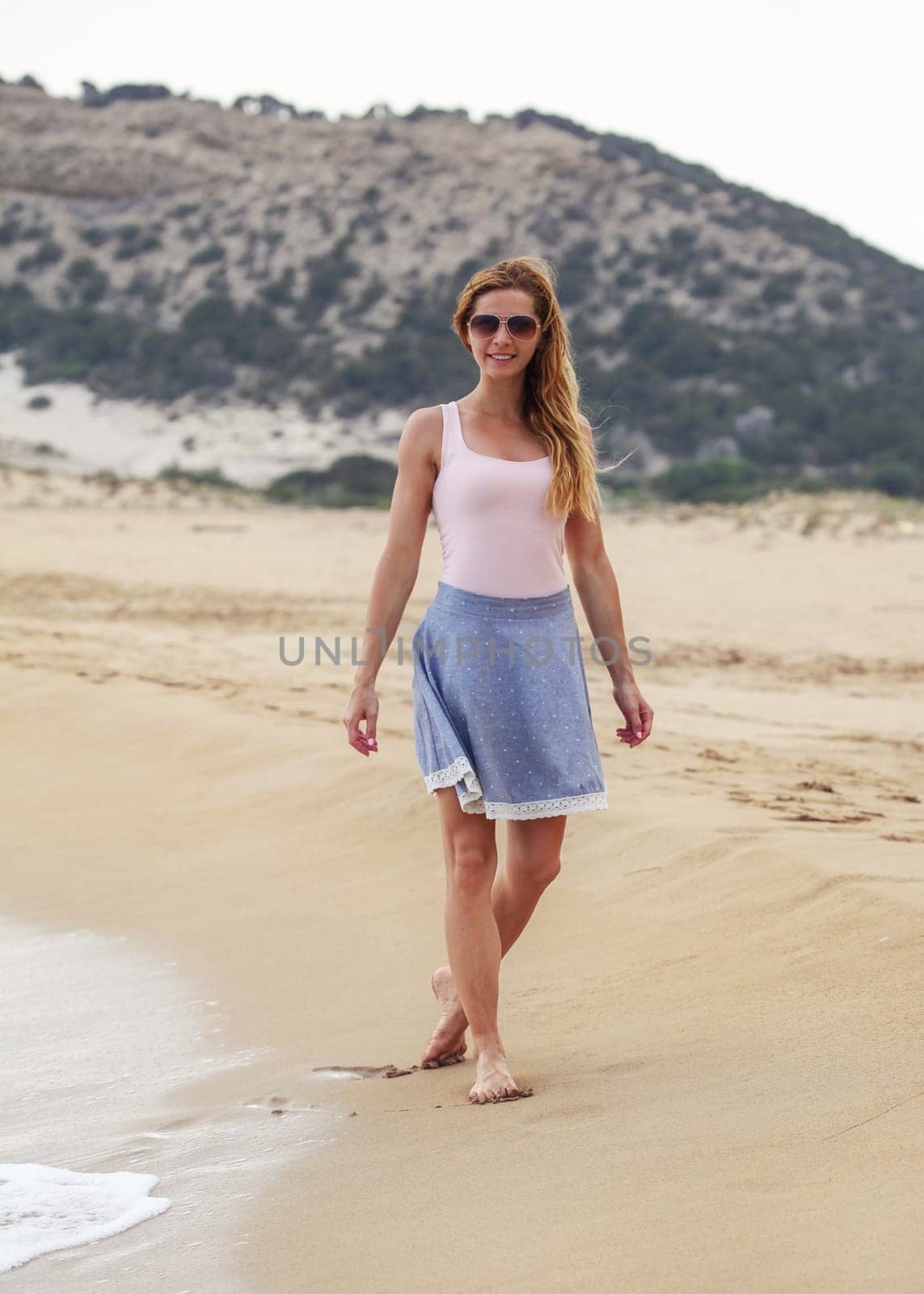 Young woman in blue skirt, t shirt and sunglasses walking on beach, smiling, overcast weather, hill behind her. by Ivanko