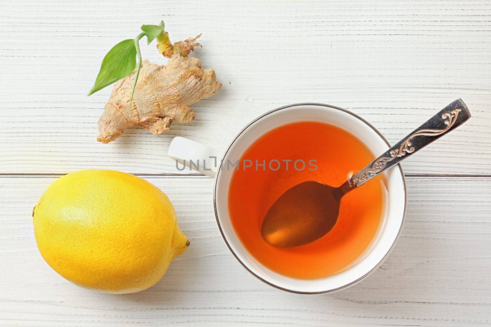 White porcelain cup, silver spoon, hot amber tea in, lemon and dry ginger root with green sprout next to it. Top view, wooden boards under.