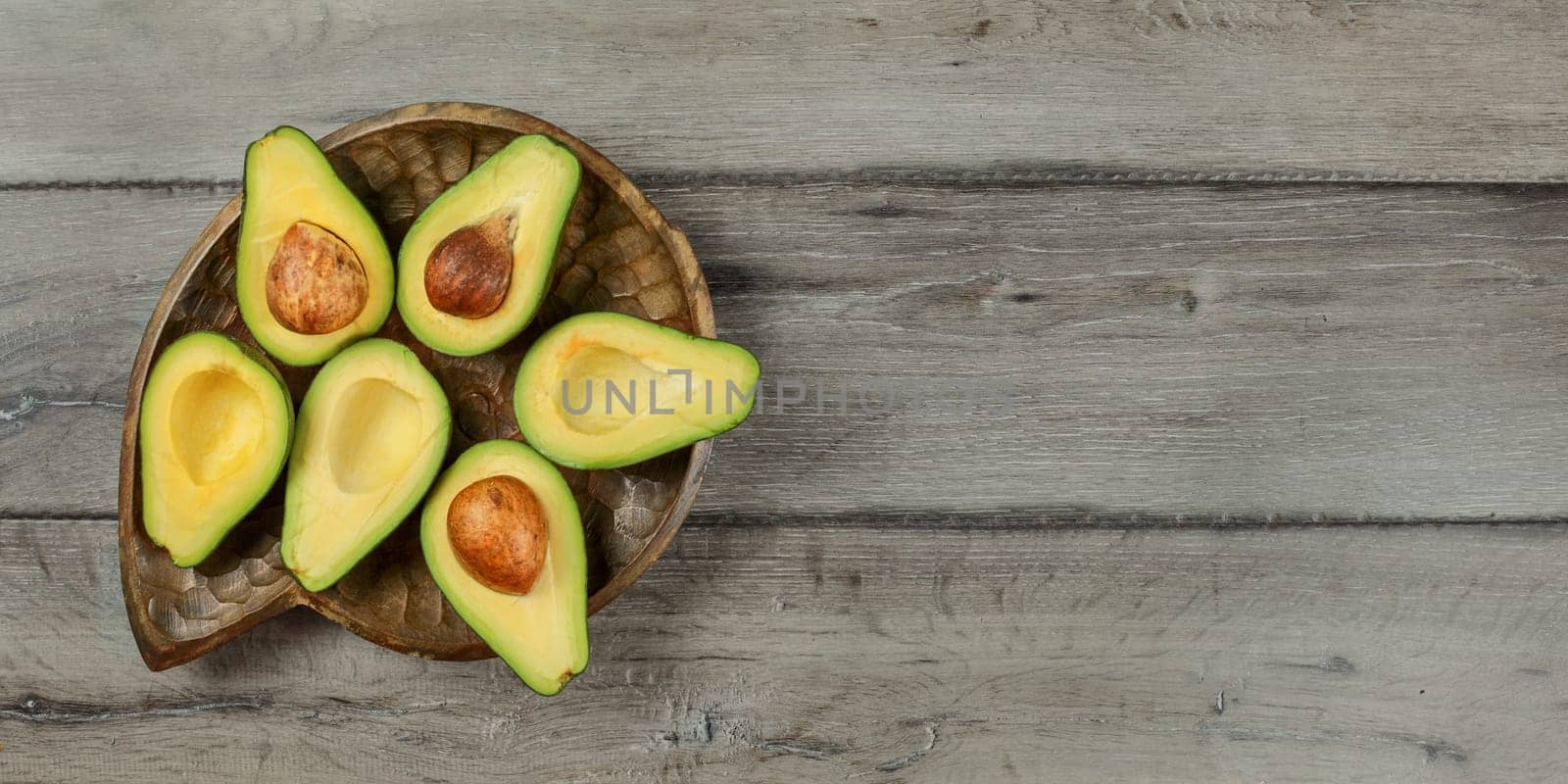Top down view, Avocados cut in half, seed visible placed on old wooden carved bowl and gray wood desk. Space for text on right.