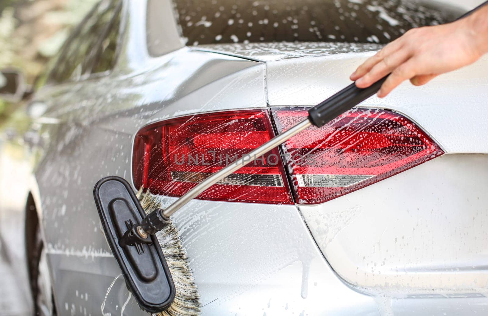 Rear light and back of silver car washed in self serve manual carwash. Brush cleaning shampoo covered surface.