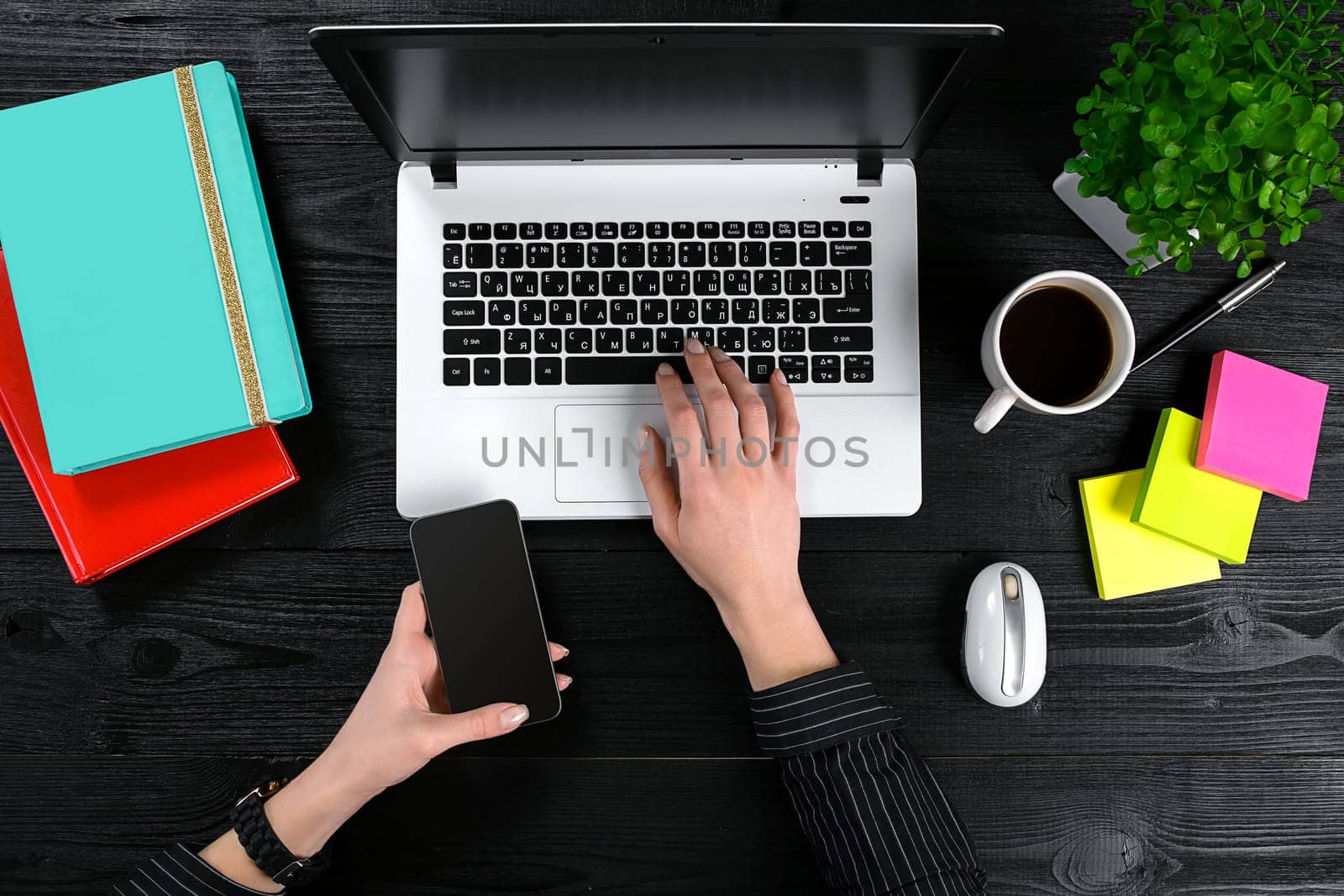 Female hands holding a smart and typing on the keyboard of a laptop on black wooden table. Mock-up with laptop, cup and notebook