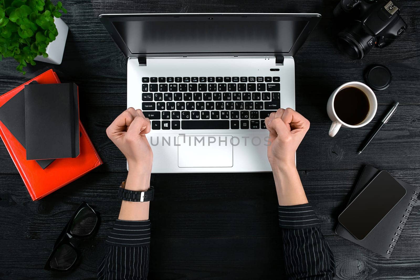 Woman working at the office table. Top view of human hands, laptop keyboard, a cup of coffee, smartphone, notebook and a flower on a wooden table background. Woman clenching her fists