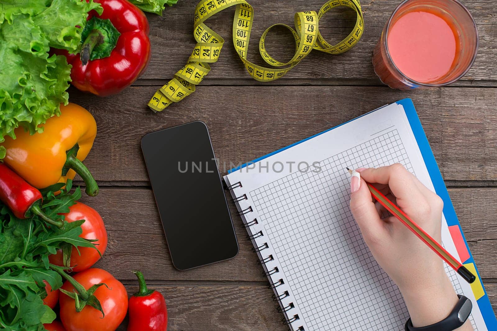 oncept diet, slimming plan with vegetables top view mock up. A female hand in a sports bracelet writes an article about proper nutrition