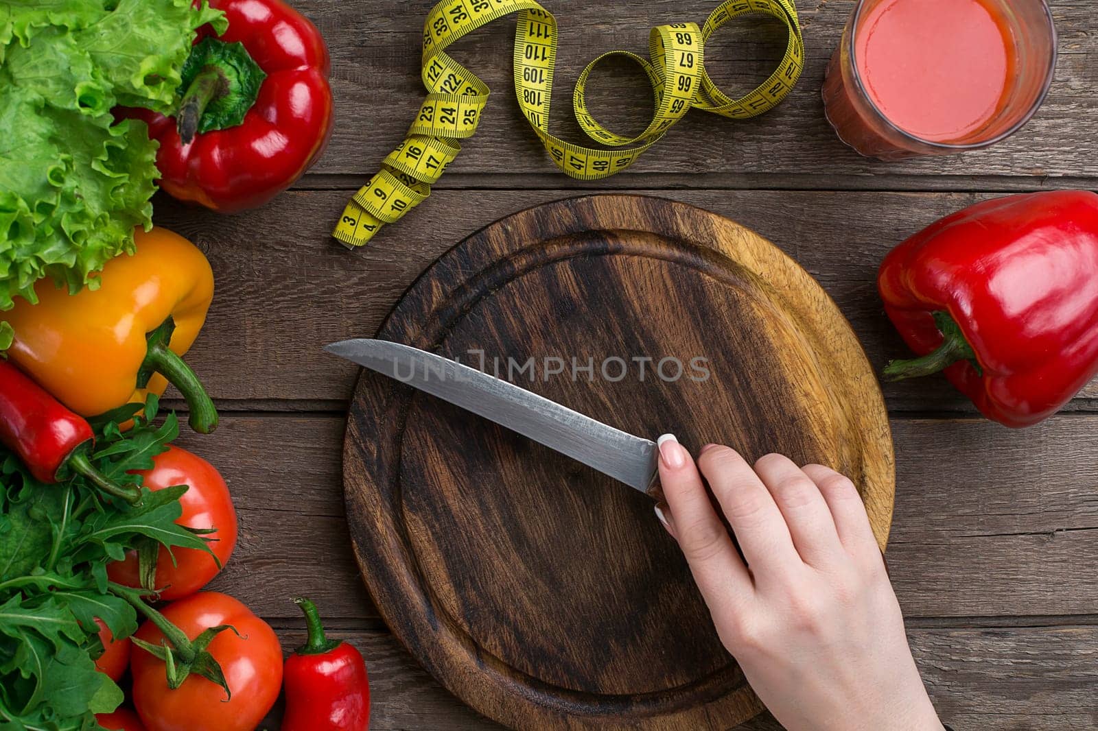 Female hands cutting vegetables at table, top view. On the table leaves of lettuce, pepper, a glass of tomato juice, a wooden board and a knife