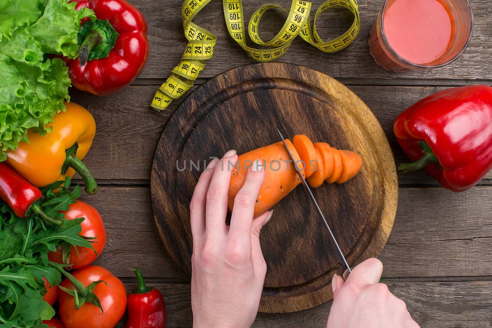 Female hands cutting carrot at table, top view. On the table leaves of lettuce, pepper, a glass of tomato juice, a wooden board and a knife