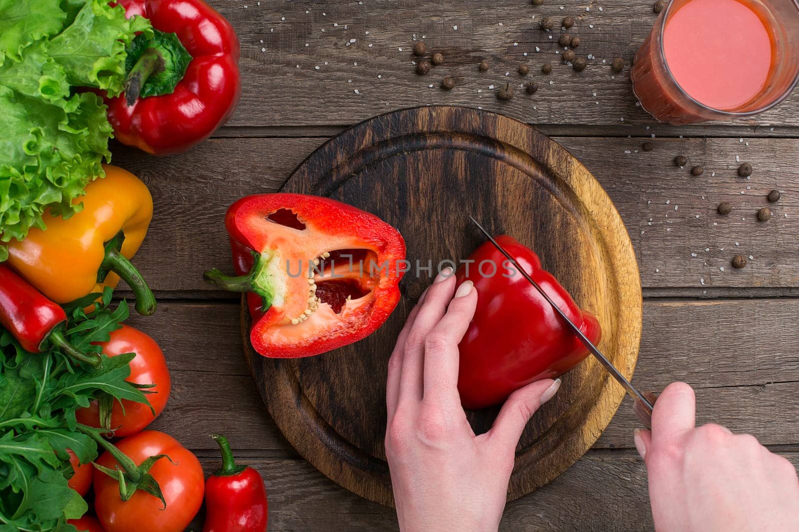 Female hands cutting pepper at table, top view. On the table leaves of lettuce, pepper, a glass of tomato juice, a wooden board and a knife