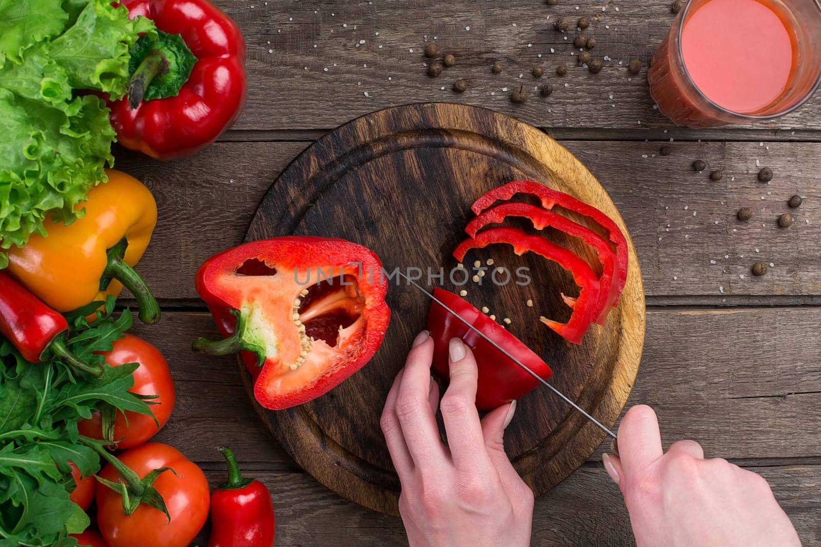 Female hands cutting pepper at table, top view. On the table leaves of lettuce, pepper, a glass of tomato juice, a wooden board and a knife