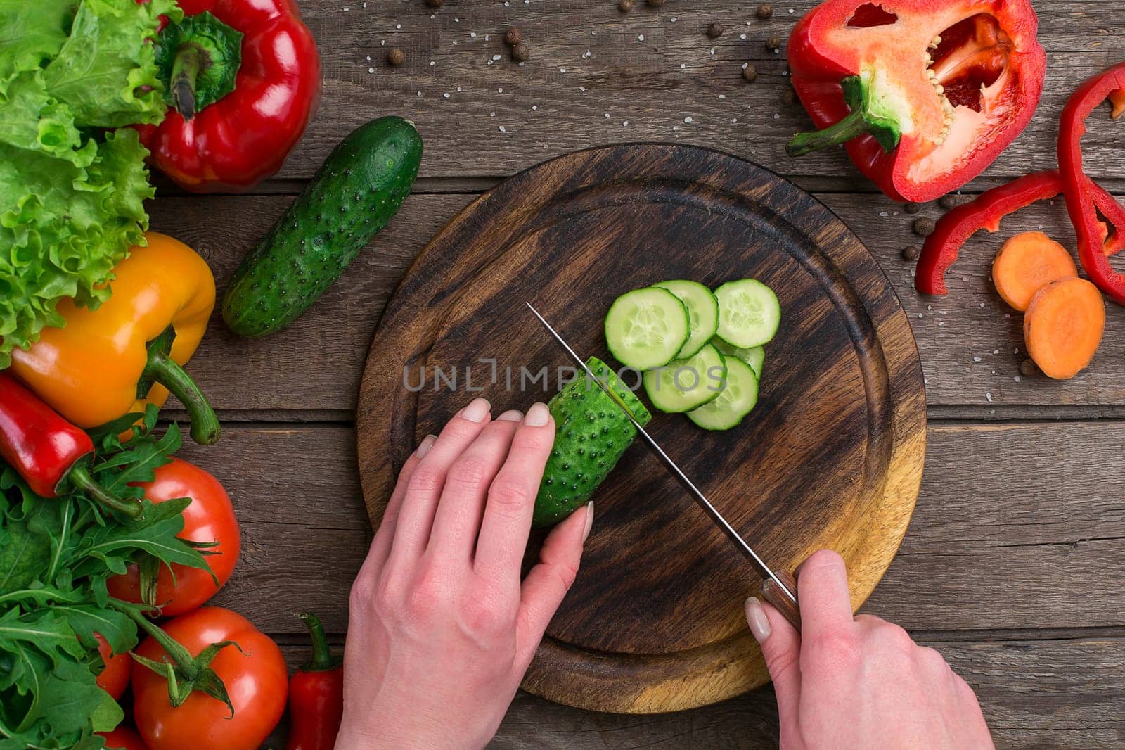 Female hands cutting cucumber at table, top view. On the table vegetables and a wooden board