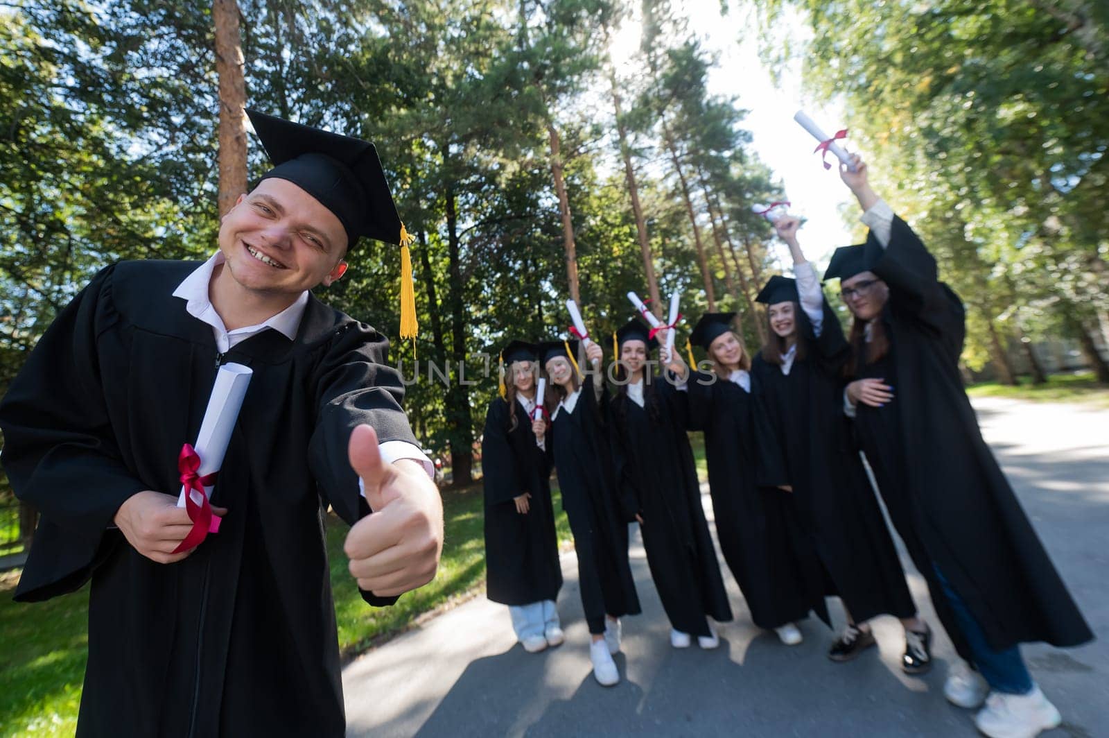 Happy young caucasian male graduate showing thumbs up. A group of graduate students outdoors. by mrwed54