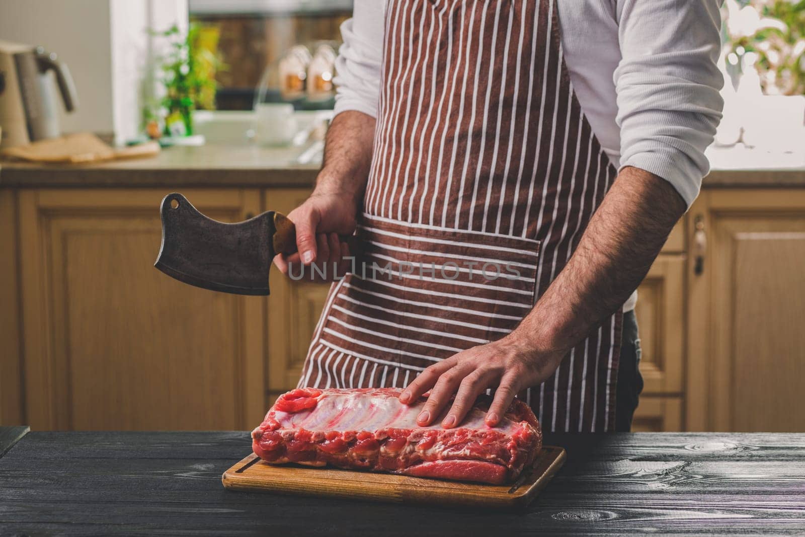 Man cuts of fresh piece of beef on a wooden cutting board in the home kitchen. A man in a striped apron with a big knife in his hands