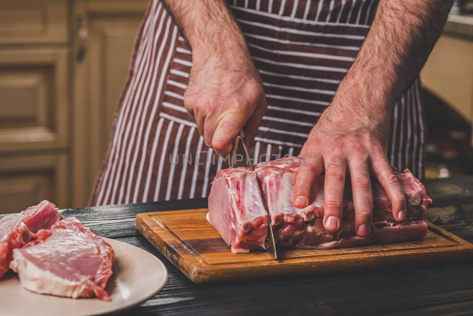 Man cuts of fresh piece of meat on a wooden cutting board in the home kitchen. A man in a striped apron with a big knife in his hands