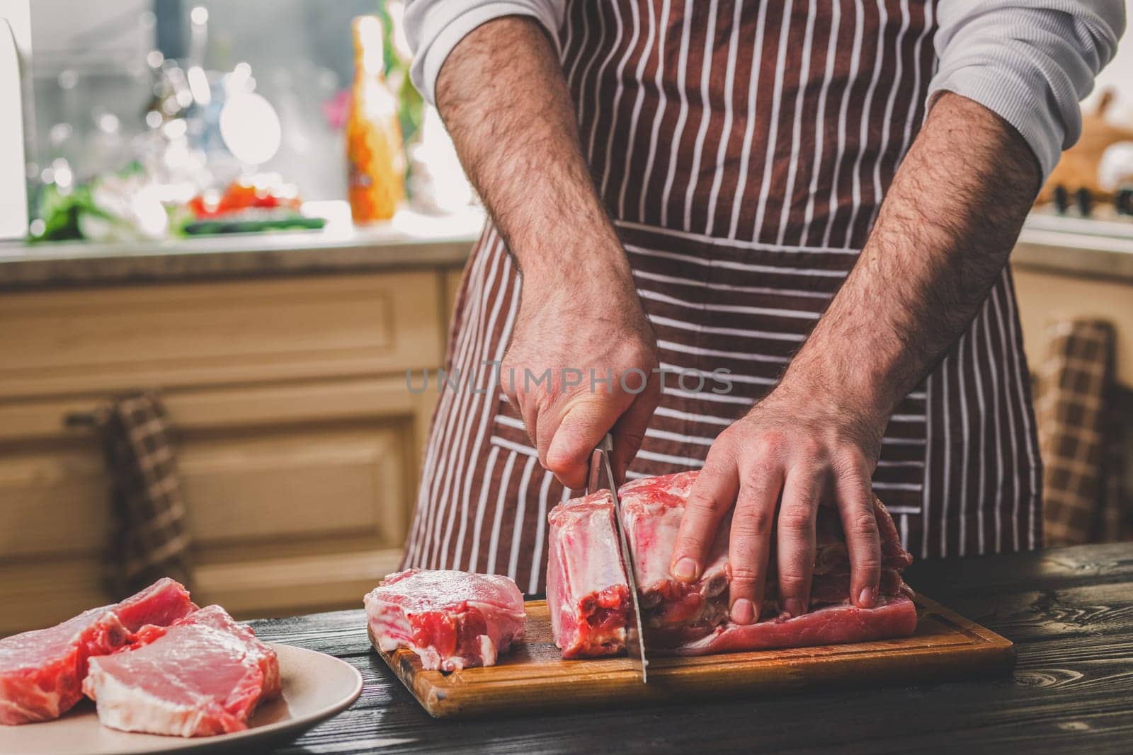 Man cuts of fresh piece of beef on a wooden cutting board in the home kitchen. A man in a striped apron with a big knife in his hands