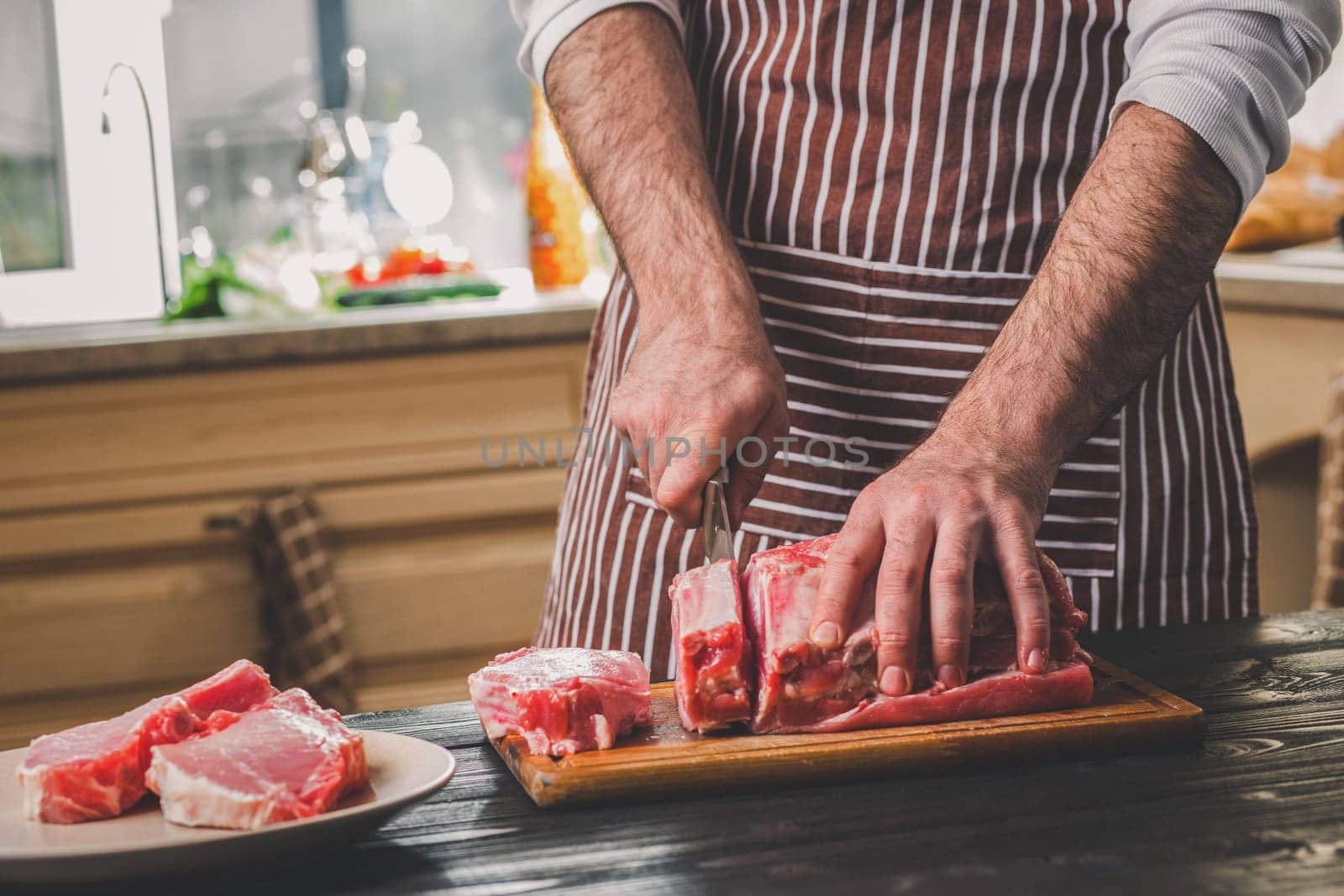Man cuts of fresh piece of beef on a wooden cutting board in the home kitchen. A man in a striped apron with a big knife in his hands