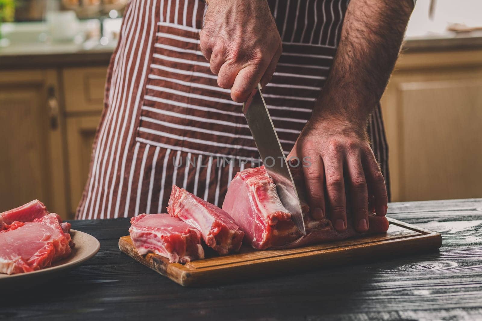Man cuts of fresh piece of meat on a wooden cutting board in the home kitchen. A man in a striped apron with a big knife in his hands