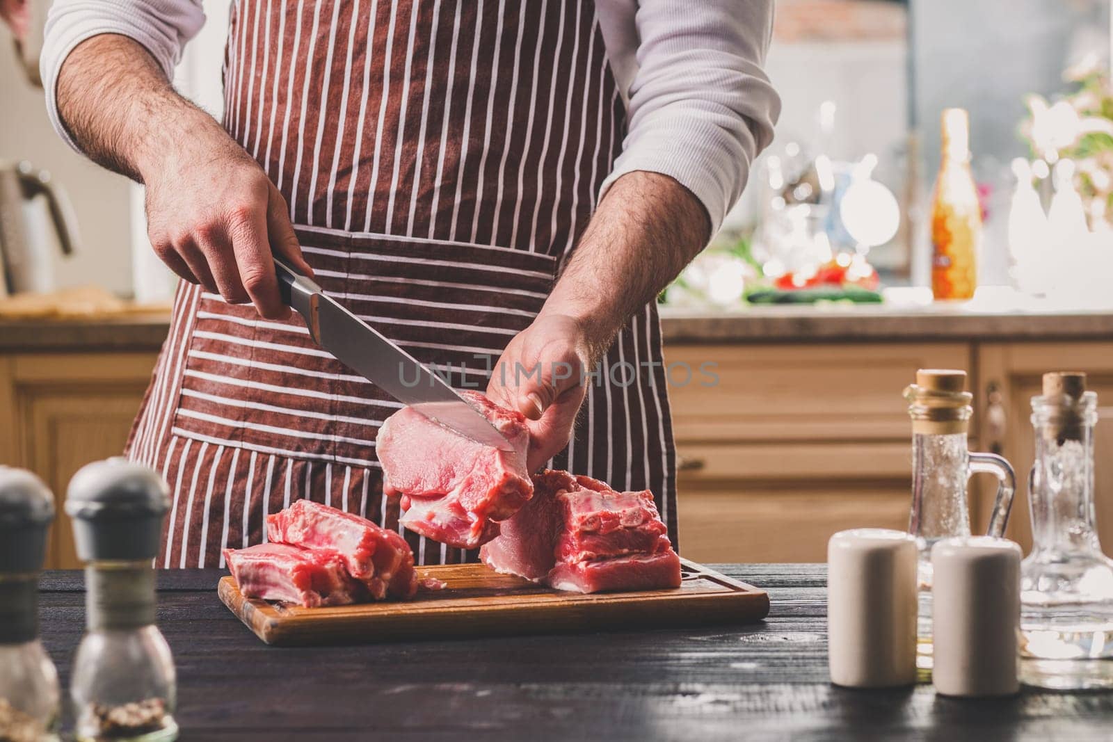Man cuts of fresh piece of meat on a wooden cutting board in the home kitchen. A man in a striped apron with a big knife in his hands