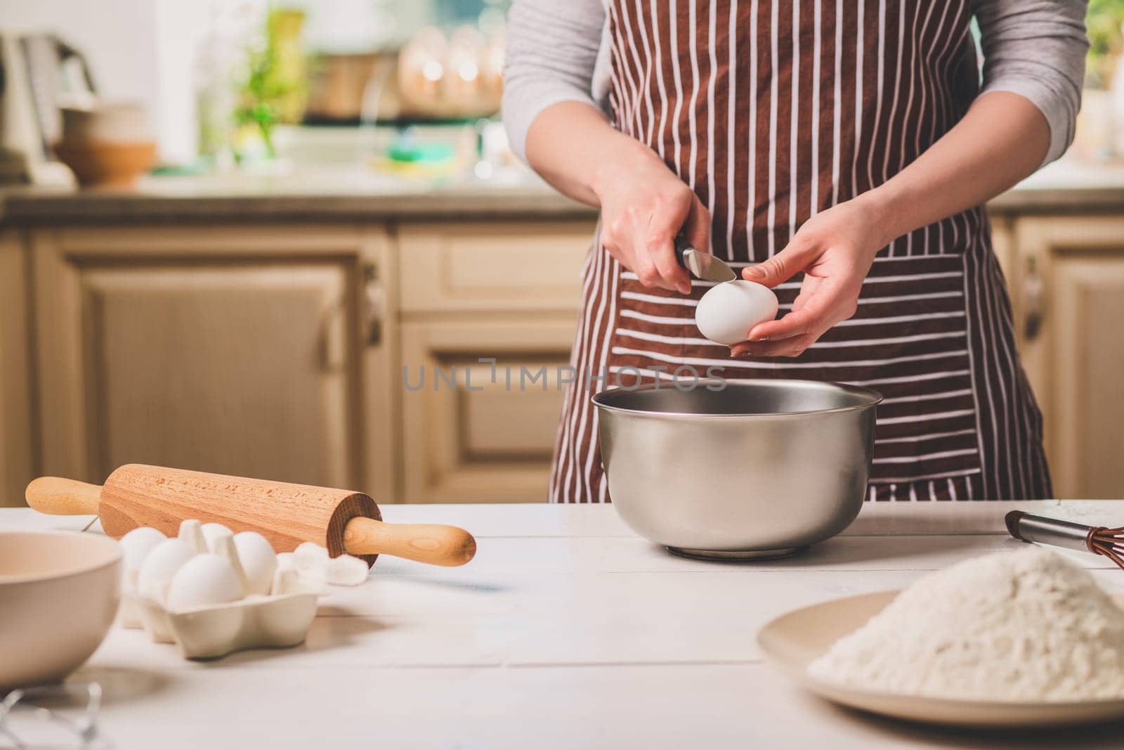 Young woman breaking egg over bowl with dough, close-up. A woman in a striped apron is cooking in the kitchen