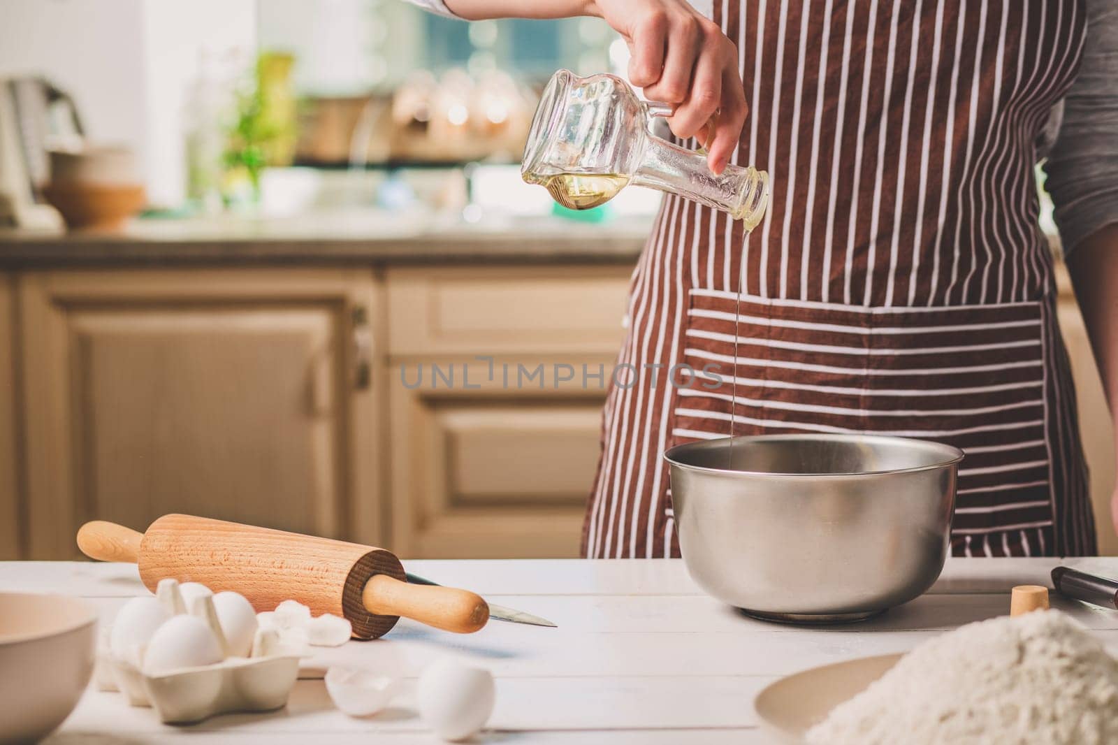 Young woman pouring oil into a bowl with dough, close-up. A woman in a striped apron is cooking in the kitchen
