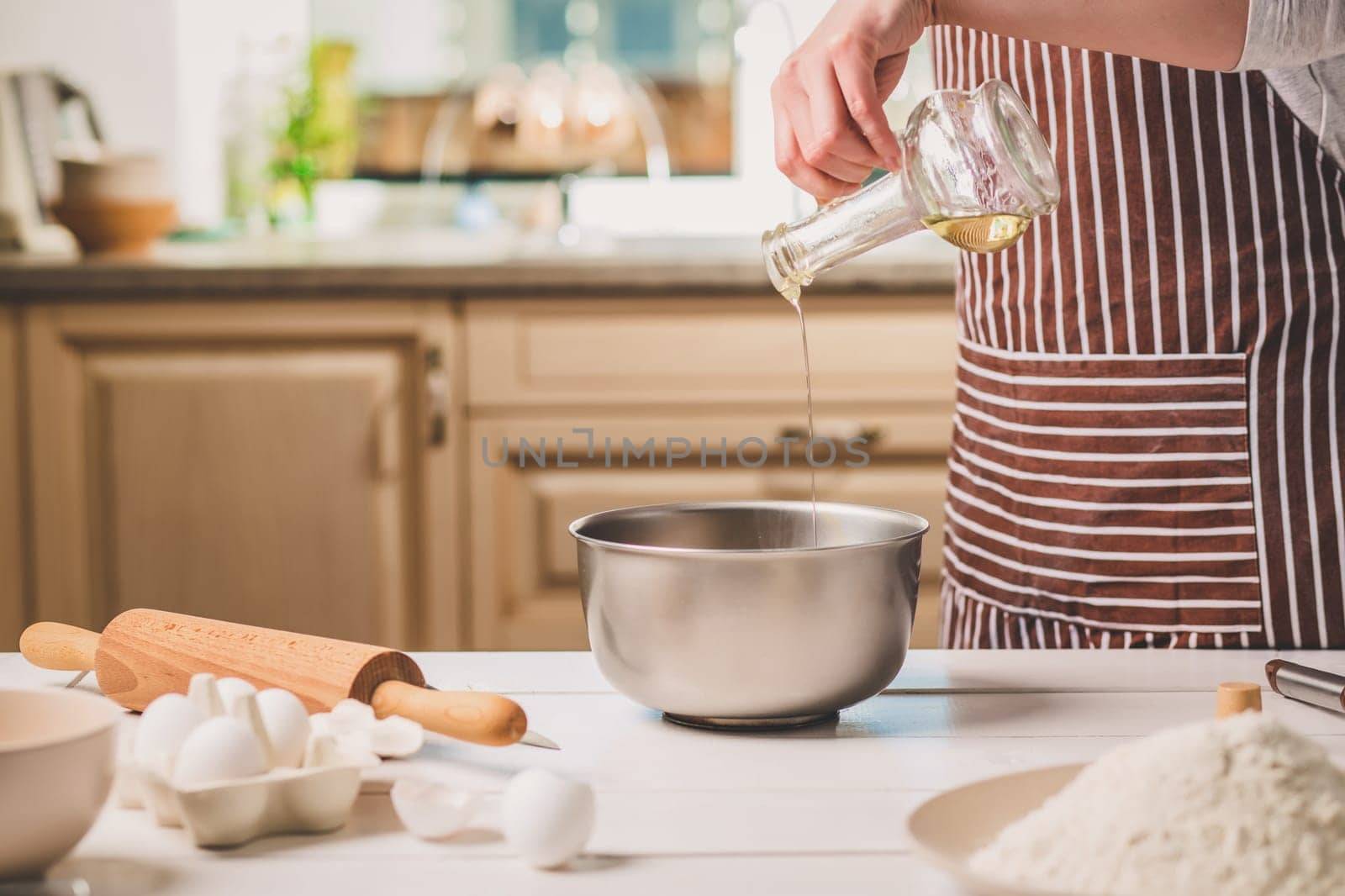 Young woman pouring oil into a bowl with dough, close-up. A woman in a striped apron is cooking in the kitchen