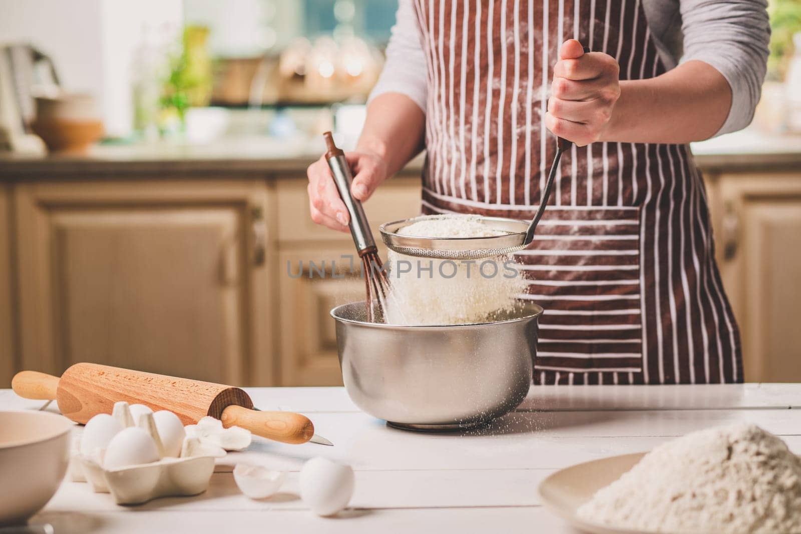Young woman holding bowl with dough and whisk, closeup. A woman in a striped apron is cooking in the kitchen