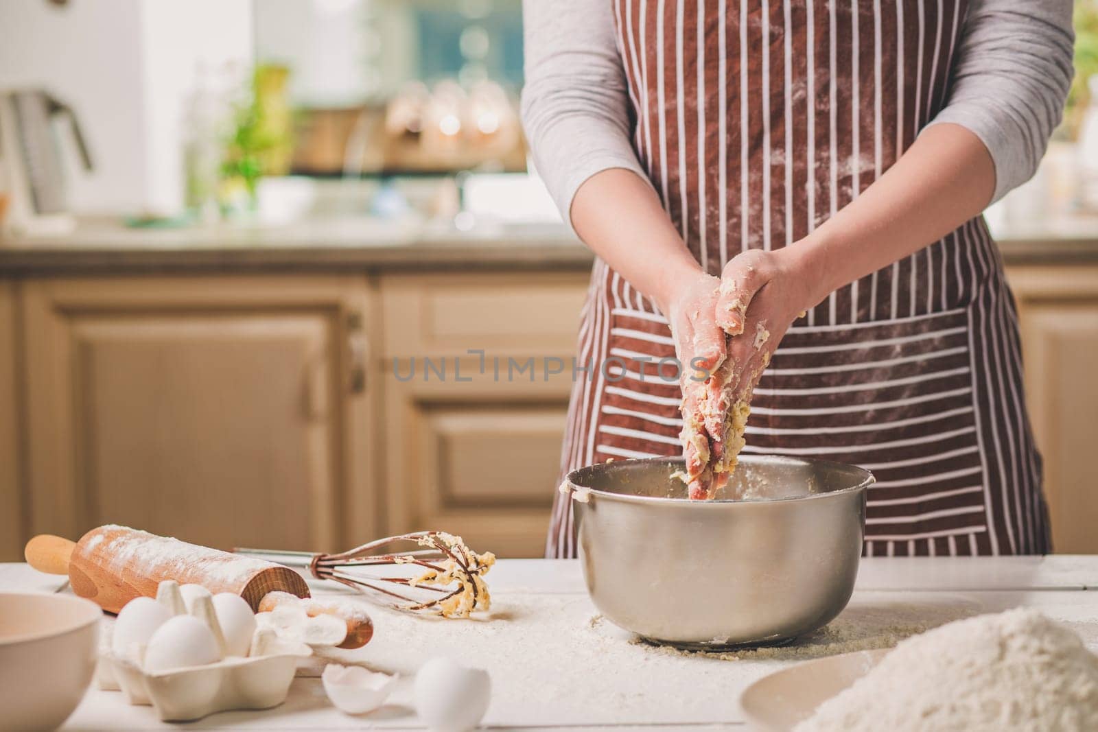 Woman hands kneading dough in iron bowl. A woman in a striped apron is cooking in the kitchen